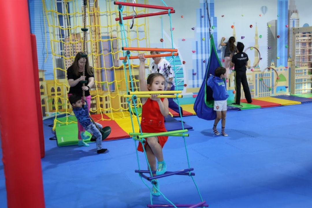 Children play in a colorful indoor sensory gym with padded flooring, climbing structures, rope ladders, swings, and a small climbing wall. One child climbs a rope ladder in the foreground while others explore equipment nearby, with adults supporting children in the background.
