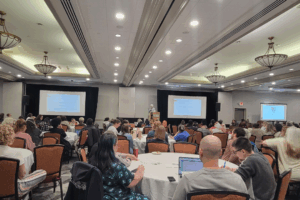 A wide view of a large conference room filled with attendees seated at round tables, listening to a speaker at a podium. Two large projection screens display presentation slides at the front of the room, and participants take notes on paper and laptops during the session.