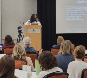 A presenter stands at a podium in a conference room, speaking into a microphone while gesturing with one hand. A large screen behind them displays a slide titled “Using Stories to Advocate Language,” with bullet points. Audience members sit at round tables facing the stage, listening.