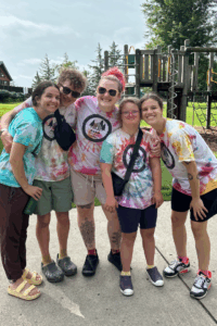 Five smiling young people wearing matching tie-dye AuSM Camps T-shirts stand close together with their arms around each other on a paved path at a playground. Behind them are green grass, trees, and playground equipment including climbing structures and slides on a sunny day.