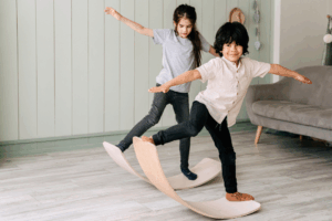 Two children play together indoors, each standing with arms outstretched for balance on curved wooden balance boards. The child in front, smiling, wears a light short-sleeve button-up shirt and dark pants. The child behind wears a grey T-shirt and dark pants, focused on balancing. They are in a bright room with light wood floors, a grey couch, and pale green paneled walls.