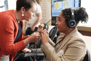 A close-up at an indoor event shows two people interacting at a booth. One person, wearing headphones, sits facing another person who is leaning in and helping adjust a small device connected by wires. Other attendees sit in the background, one wearing tinted glasses, with a screen behind them displaying colorful brain-style graphics.
