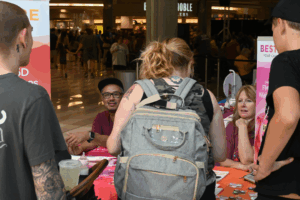 A crowded indoor public space with a resource table in the foreground. Several adults stand around the table talking with staff seated behind it, which is covered with pink materials, flyers, and small items. A large crowd moves through the open area behind them, creating a busy, energetic event atmosphere.