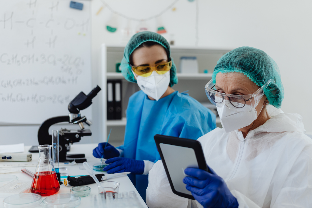 Two researchers wearing protective lab gear, including masks, gloves, and hair covers, work at a laboratory bench. One person looks at a tablet while the other prepares a sample near a microscope, with lab glassware and equipment visible on the table.