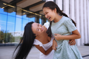 An adult and a child stand outdoors in front of a modern building, laughing together as the adult gently lifts or tickles the child. Both appear joyful and engaged in a playful moment.