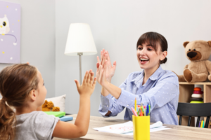 An adult and a young child sit across from each other at a table, smiling and giving each other a high five. Art supplies and toys are visible, suggesting a supportive learning or therapy activity.