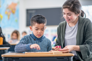 An adult works one-on-one with a young child at a classroom desk, guiding the child as they manipulate a hands-on learning material. Other students and classroom elements are visible in the background, indicating an inclusive learning environment.