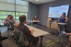 A small group of adults sit at tables in a modern meeting room, listening to a presenter standing at the front. The presenter gestures while speaking beside a table with a laptop, and a large screen behind them displays a slide titled “Ooïds” with an image of white, round shapes. Attendees face the screen, some taking notes, while large windows along one wall let in natural light and show greenery outside.