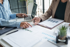 Two people sit at a desk reviewing paperwork together. One person points to a printed document with charts and graphs, while the other holds a pen and gestures toward the page. A laptop, clipboard, and additional documents are spread across the table, suggesting collaborative planning or problem-solving.