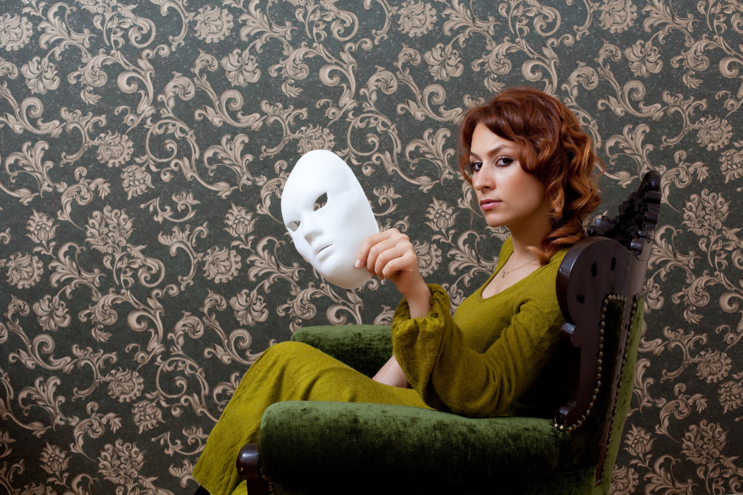 A person sits in a green upholstered chair against a patterned wallpaper background, holding a white theatrical-style mask slightly away from their face. They wear a green dress and look directly toward the camera, creating a visual contrast between the mask and their uncovered expression.