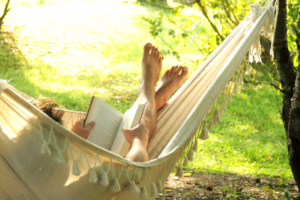 A person reclines in a fabric hammock outdoors, holding an open book while resting their bare feet toward the sunlight. The hammock is strung between trees in a green, leafy setting, creating a calm and restful scene.