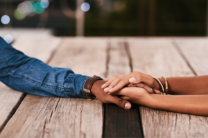 Two people sit across from each other at a wooden table, gently holding hands in the center of the frame. One person wears a denim shirt and a watch, while the other wears bracelets. The image focuses on the hands, conveying connection and support, with the background softly blurred.