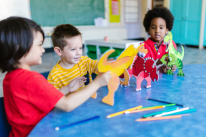 Three young children sit at a classroom table, playing together with colorful dinosaur cutouts. One child holds up a dinosaur figure while the others look on and interact. Colored pencils rest on the table, and the classroom setting is softly visible in the background, suggesting collaborative play and learning.