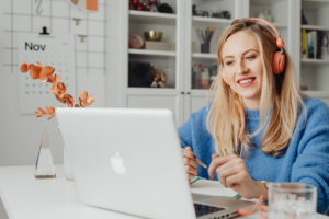 A person sits at a desk using a laptop while wearing over-ear headphones and smiling. They hold a pen and appear to be engaged in an online meeting or focused work. The setting looks like a home office, with shelves, a calendar on the wall, and a small vase with dried flowers in the background.