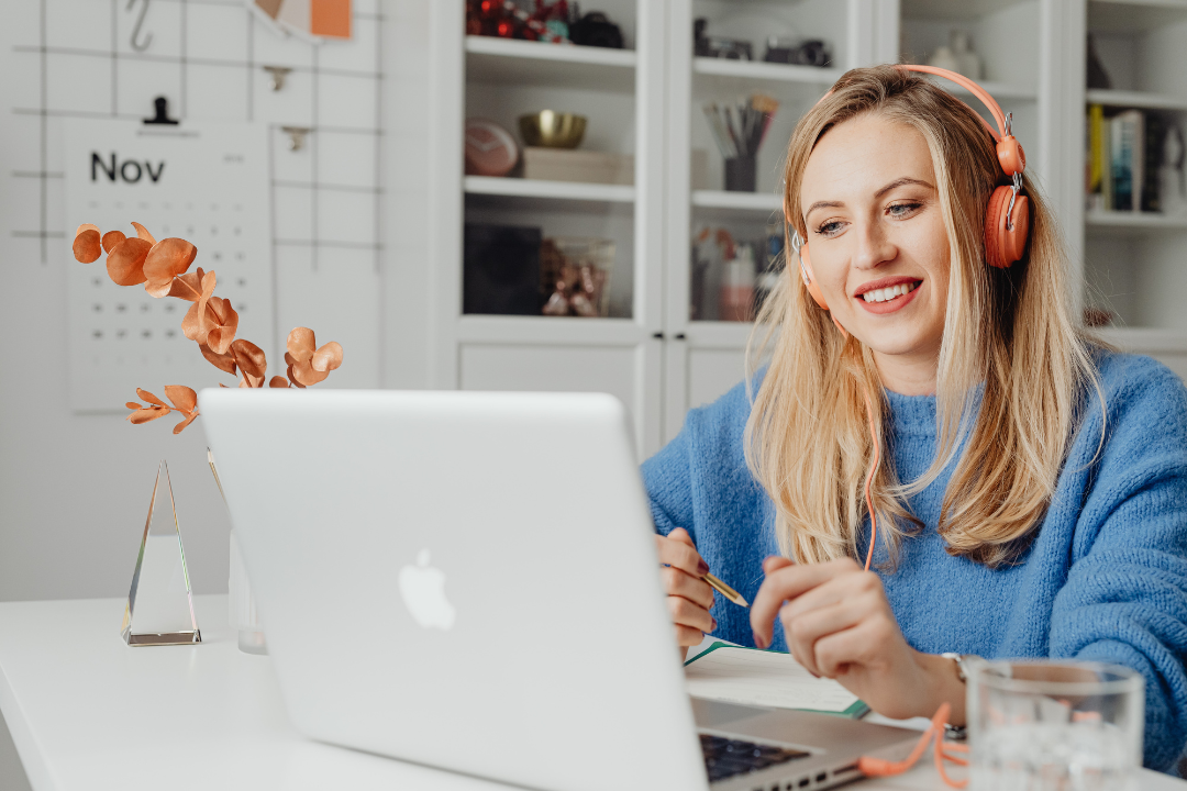 A person sits at a desk using a laptop while wearing over-ear headphones and smiling. They hold a pen and appear to be engaged in an online meeting or focused work. The setting looks like a home office, with shelves, a calendar on the wall, and a small vase with dried flowers in the background.