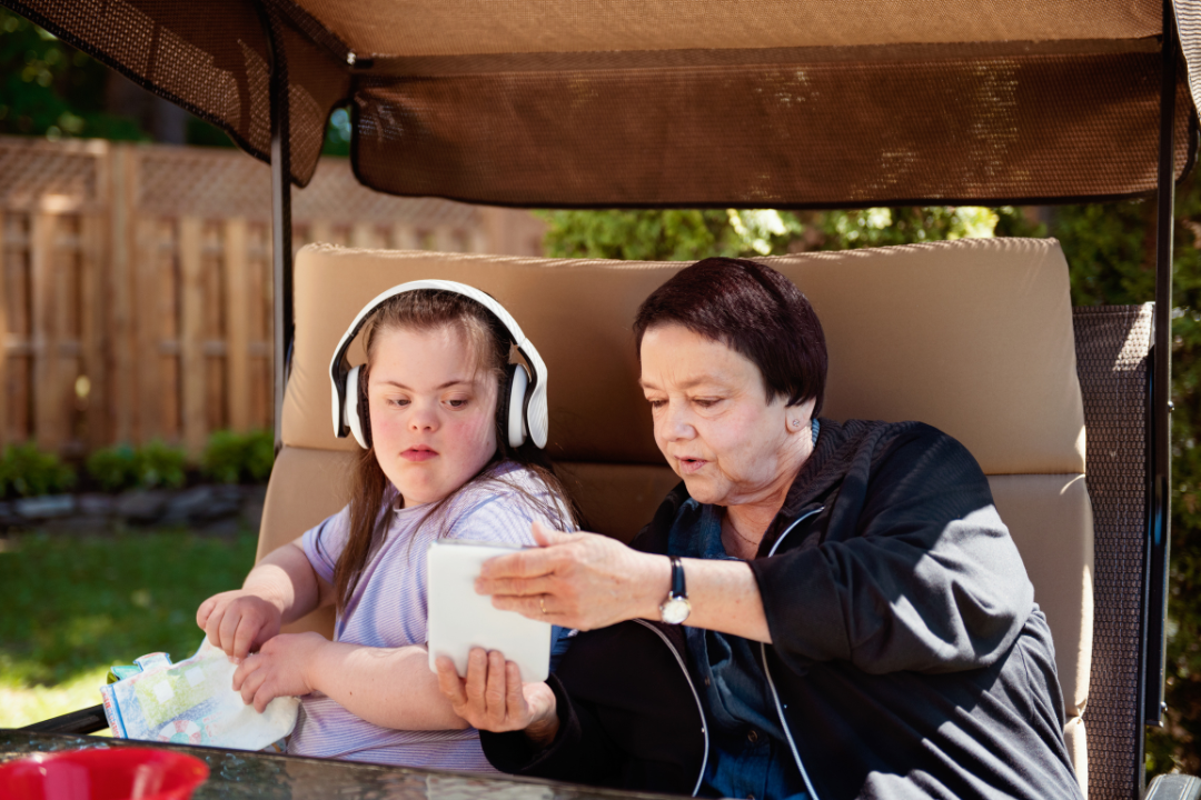 An older woman sits outdoors on a cushioned swing seat beside a young person wearing over-ear headphones. The older woman holds up a small white card or booklet and points to it, while the young person looks on and holds a patterned cloth. A table and a fenced backyard are visible in the background.