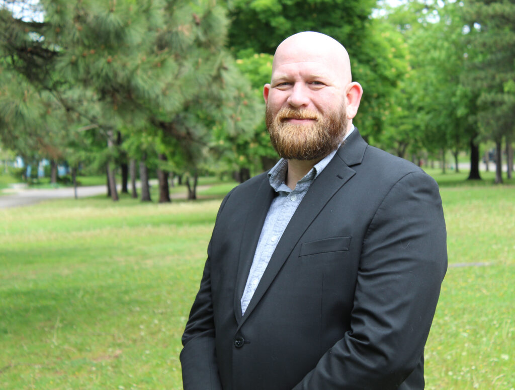 A white man with a dark blonde beard dressed in a black suit holding his hands in front of his body standing in front of a greenery background