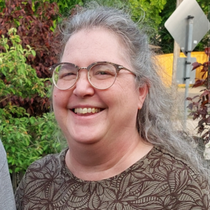 AJ Hokland - A white non-binary person with glasses and long gray hair who is smiling.
